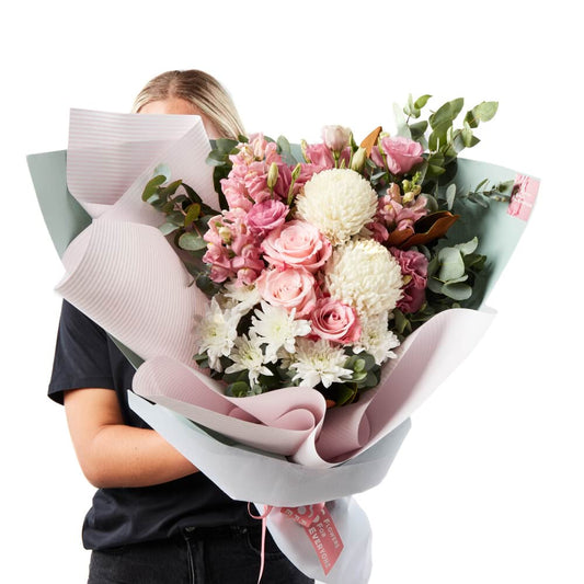 A person holding a soft pastel bouquet of various flowers including roses and eucalyptus, with pink and green wrapping.